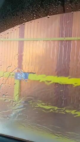 A video showing a car wash from the perspective of someone inside a vehicle. The camera focuses on the interior wall of the car wash, which is made of corrugated metal panels in shades of orange and brown. A yellow metal frame runs horizontally and vertically along the wall. A blue sign with white text reading "ВОДА" (water) and "ВОСК" (wax) is attached to the yellow frame. A silver nozzle, likely from a spray wand, is visible in the lower left portion of the frame, angled towards the wall. Water is visibly spraying from the nozzle and running down the wall, creating streaks and water droplets on a transparent barrier in front of the camera. The mirror of the vehicle is visible in the bottom left of the frame, also covered in water droplets. The scene suggests the vehicle is in the process of being washed.