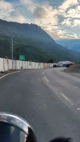 The video shows a point-of-view shot from a vehicle driving on a winding road. The road curves to the right, with a white painted line marking the edge of the lane. On the left side of the road is a low, white concrete wall topped with a fence. Beyond the wall, there are some green signs and a tall pole. Further along the road, several buildings are visible, some with red tents set up. Parked in front of the buildings are a few cars and a truck. The landscape is mountainous, with steep, green slopes and snow-capped peaks in the distance under a cloudy sky. The overall impression is of a scenic mountain road, possibly in a tourist area.