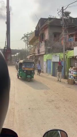 The video shows a street scene with several vehicles and buildings. In the foreground, a person is riding a motorcycle, with the rear of the motorcycle seat visible. On the road, there is a green auto-rickshaw with people inside, followed by several motorcycles. A large construction crane is visible in the background, along with ongoing construction work. Buildings with multiple floors line the side of the street, some with balconies and clothes hanging out to dry. A few people are walking along the sidewalk. The road appears to be unpaved and dusty, suggesting a rural or developing area. The sky is partly cloudy. The overall atmosphere is active and suggests a busy street with both daily traffic and construction activities.