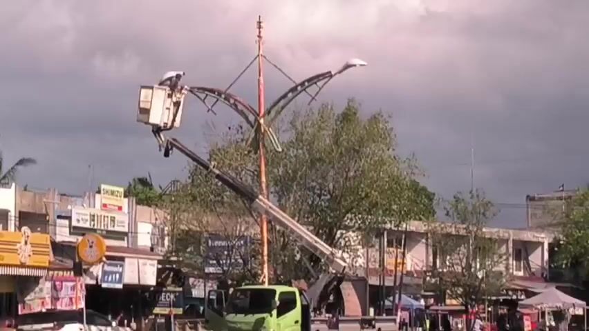 The video shows a worker in a bucket lift repairing or maintaining a tall street light fixture. The light is a decorative structure with curved metal arms supporting multiple light sources. The lift is mounted on a green truck. The background includes commercial buildings with signs in a somewhat urban environment under a cloudy sky.