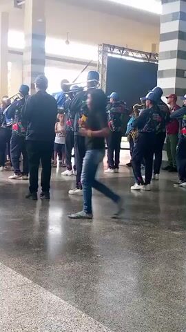 A group of people in matching uniforms, some holding musical instruments, are gathered indoors. One person in the foreground, wearing a dark jacket and blue jeans, walks across the frame. The group appears to be a marching band or ensemble, with some members playing instruments like trumpets and trombones. They are standing on a polished concrete floor, and in the background, there is a stage with a dark screen and some lighting equipment. The environment appears to be a large indoor space with columns and natural light coming from the left. The individuals are mostly facing forward or towards each other, suggesting they are either performing or preparing to perform.