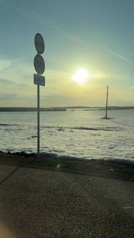 The video pans across a snowy field with a sunlit sky. The sun is bright in the distance. There are some trees, a road, a guard rail, and a utility pole visible in the shot. There is a sign on a pole at one point in the pan.