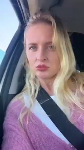 A woman with long blonde hair is sitting in the driver's seat of a car. She's wearing a pink cardigan over a white top and a seatbelt. The car is moving, as evidenced by the blur of the background visible through the side window. The woman's hair is being blown around her face, presumably by the wind. Her expression is somewhat neutral or perhaps slightly annoyed.