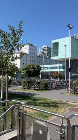 A sunny daytime exterior scene shows a large building with a modern, light blue facade and a glass-covered entrance. In front of the building is a massive bicycle parking area filled with hundreds of bicycles. Trees and bushes are planted between the building and a paved area with a sidewalk. A few individual bicycles are parked near the bushes. A motorcycle is moving on the paved area towards the building. A bus and several cars are parked on the street in front of the building. A person on a scooter is also on the street. Traffic signs are visible.