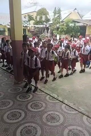 The video shows a group of young students, mostly boys, in school uniforms, walking in a line. They are wearing white shirts, red shorts, and some have red caps. Many of them are carrying backpacks. The students appear to be walking out of a building or into an open area, possibly a schoolyard. The environment looks like a school, with a building in the background and some trees. The time is daytime, as indicated by the lighting. Some adults, possibly teachers, are also visible among the students. The students are walking in an orderly fashion, and some are looking at the camera. The focus of the video is on the students and their movement.