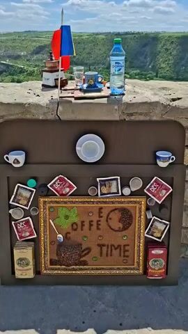 The video shows a coffee setup with a scenic view. On a stone ledge, there is a coffee grinder, a small cup with an Italian flag and a Moldovan flag attached, a glass of water, a cup of coffee with a saucer, a croissant, and a bottle of water. Below the ledge, there is a display of coffee-related items, including coffee cups, coffee pods, and framed pictures. The background features a lush green landscape with hills and a valley under a bright, cloudy sky.