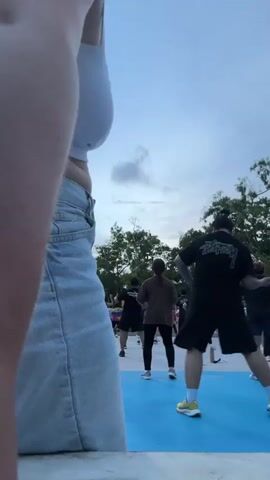 The media shows an outdoor dance or exercise class in Phuket City Municipality, Thailand. The scene is set on a bright blue court, possibly a basketball or multipurpose court, with a white border. In the foreground, a woman in a white crop top and denim shorts is dancing dynamically. She is facing away from the camera, then turns to face it as she moves. Her arms are raised and her expression is energetic.

In the background, a group of people, likely participants in the class, are also dancing or exercising. They are positioned behind the woman in the foreground. Most of them are facing away from the camera. Some are wearing bright pink tops, while others are in darker clothing. The time of day appears to be dusk or early evening, as the sky is a pale blue with some clouds, and a single bright light fixture is visible on the left side of the frame. Trees with lush green foliage are visible in the background, suggesting a tropical location.

The overall mood of the video is lively and active, with a sense of community as people participate in a group activity outdoors. The weather seems pleasant for an outdoor activity. There is no visible text in the media file.