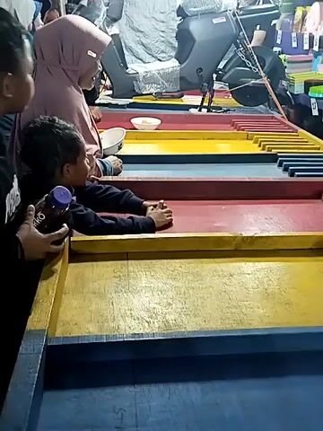 A low-angle medium shot shows an adult woman wearing a hijab and two boys (all viewed from the side) participating in a ball game at what appears to be an outdoor fair or market at night. The game involves aiming and rolling small yellow balls into designated slots. The game table has multi-colored horizontal sections - blue, yellow, red, yellow. Behind them, an electric scooter is on display, possibly as a prize. A person is seen operating the game from behind the scooter. A variety of household items, such as storage containers, are displayed on a shelf. There are several people moving in the background.