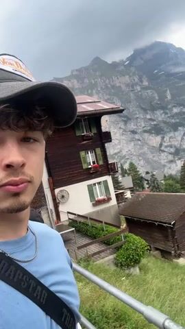 A person wearing a baseball cap and a light blue shirt is standing in front of a picturesque Swiss village. The village is nestled in a valley with traditional wooden houses with green shutters. In the background, majestic mountains with snow-capped peaks loom against a cloudy sky. The camera is held at a slight angle, capturing a selfie with the stunning mountain landscape as the backdrop. The person is looking towards the camera, with a serene expression. The video appears to be taken from a moving vehicle or a viewpoint overlooking the village, as indicated by the slight motion blur and the railing in the foreground. The overall scene evokes a sense of tranquility and natural beauty.