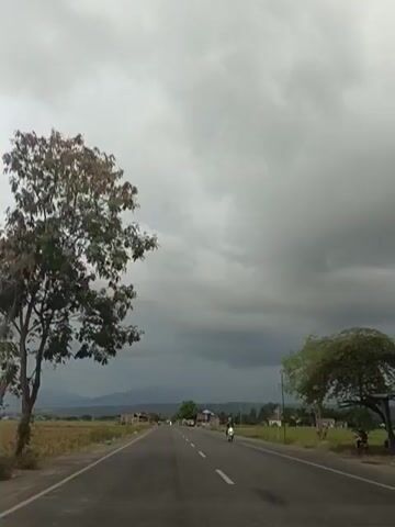 A video shows a paved road stretching into the distance, flanked by fields and trees. The sky is overcast with dark, heavy clouds, suggesting impending rain or a storm. A lone motorcycle is seen traveling on the road. The overall atmosphere is somewhat somber due to the weather conditions.