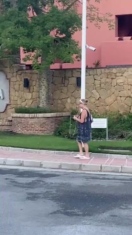 This video captures a woman walking on a sidewalk in front of a building with a sign that reads "TORRE BERMEJA" with four stars beneath it. The building appears to be an apartment complex or hotel, with a coral-colored facade and balconies visible. The architecture suggests a warm climate, possibly Mediterranean, consistent with the provided location context of Resinera-voladilla, Spain.

The woman is wearing a dark-patterned dress and flip-flops, carrying a phone and a bag. She seems relaxed as she walks, looking down at her phone. The scene appears to be outdoors during the daytime, with lush green trees and landscaping surrounding the building and sidewalk. The lighting suggests it might be late afternoon or early evening, as the light is soft and diffused. There's a hint of greenery and perhaps a street in the foreground, indicating this is a public area. The overall mood is tranquil and everyday.