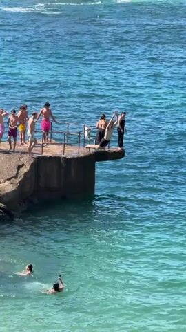 A group of young people are gathered on a concrete platform overlooking a body of turquoise water. Several individuals are standing near the edge of the platform, some leaning on a metal railing. One person is in mid-air, having just jumped from the platform into the water. Below, in the water, several other individuals are swimming. The water appears clear and the day is sunny. The scene is of recreational activity in an ocean environment.