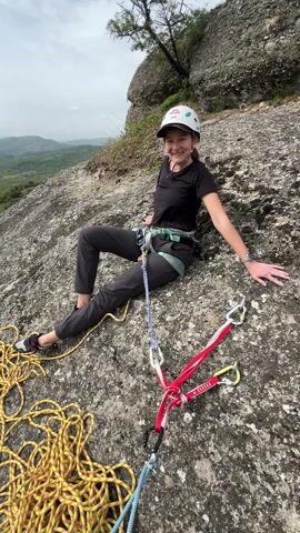 The video shows a young woman wearing a helmet and climbing harness sitting on a rocky surface, presumably at the top of a climb. A yellow rope is coiled beside her, and rock climbing equipment is nearby. She smiles at the camera. The camera pans from her to reveal a wide view of a green valley with mountains in the background. The sky is overcast.