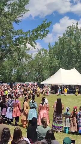 A diverse group of people are gathered outdoors in a grassy area. Many individuals are wearing traditional indigenous clothing, including colorful dresses, skirts, and beaded headdresses. Some are dancing in a central area, while others are observing the event. In the background, there are trees and a large white tent. The sky is blue with fluffy white clouds. The overall scene appears to be a cultural gathering or festival.