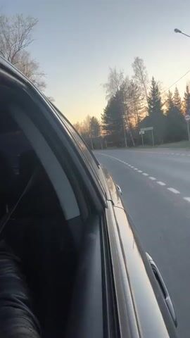 The video shows a view from inside a moving vehicle. The camera is pointed out the passenger side window. The road is straight and paved, with trees lining both sides. The sky is a clear blue with a hint of orange near the horizon, suggesting it is either sunrise or sunset. There are a few other vehicles visible on the road ahead. A portion of the car's interior is visible, including the seat and the door panel.