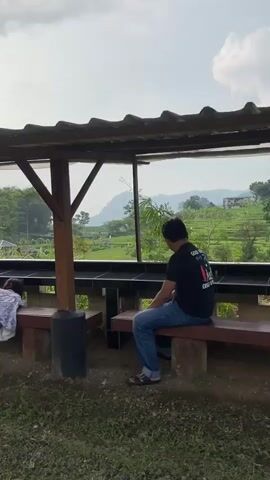 A young girl in a dress is lying on a wooden bench, her head and upper body hanging over the side, looking at the ground. A man in a black t-shirt and jeans is sitting on the same bench, looking out at the landscape. They are under a shelter with a metal roof and wooden support beams. The landscape beyond the shelter features green fields, terraces, trees, and a distant mountain. The sky is overcast.