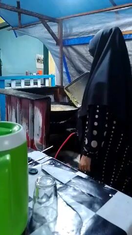A person wearing a black hijab and a black and white polka dot dress stands in front of a food stall. They are holding a metal tray over a cooking surface that appears to be a grill or stove. A green and white insulated container and a glass are on a checkered surface in the foreground. The stall has a blue tarp roof and appears to be outdoors or in an open-air setting. A red hose is connected to the cooking equipment.