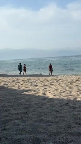 The scene is a sandy beach on a partly cloudy day, with a view of mountains across a body of water. In the distance, mountains are visible under a hazy sky. The water is calm, with small white buoys floating in the distance. The foreground is composed of sandy beach with subtle ripples and shadows, suggesting some vegetation is nearby. Three people are walking along the water's edge towards the camera. On the left, a person carries a green and white striped towel. They are accompanied by another person in a bikini, and a third person is on the right, also in a bikini. A small dog is running in the shallow water near them. The audio suggests music is playing in the background.