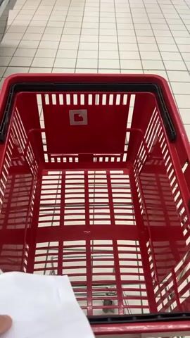 The video shows a first-person perspective of someone pushing a red plastic shopping basket through a supermarket in Saint-Galmier, France. The person's hand is visible in the foreground, holding a crumpled piece of white paper, possibly a shopping list.

The supermarket features white tiled flooring with a grid pattern. As the basket moves, various supermarket elements come into view, including refrigerated display cases, shelves stocked with products, and promotional stands.

Prominently displayed are several large cardboard boxes advertising "ARGAL" branded products, specifically "TRADITION ESPAGNOLE" (Spanish Tradition), indicating they are a Spanish-style food brand established "DEPUIS 1916" (Since 1916). Some boxes also feature the tagline "PARTAGEZ L'AUTHENTICO" (Share the authentic). Small price signs displaying "4€" and "5€" are visible near these displays.

The shopping basket itself is empty and made of red plastic, with a small red logo on its interior back panel. The sounds of the basket wheels rolling on the floor are audible throughout the clip. The store appears well-lit, suggesting daytime. No other people are clearly visible in the short video.