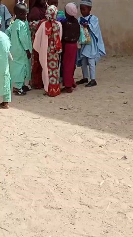 The video shows a group of children, most in traditional attire, standing on a sandy area outside a building. Some children are wearing light green robes and caps, while others have patterned dresses and headscarves. A few are carrying items like a green bucket and a white plate. One child is eating something wrapped in fabric. The environment appears to be a village or rural area with modest buildings in the background.