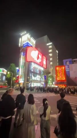At night, a low-angle shot shows a bustling crosswalk intersection in a Japanese city. Pedestrians wait on the sidewalk, ready to cross. Illuminated signs and billboards adorn the surrounding buildings, displaying advertisements and glowing symbols. The "BLUE ELEPHANT" sign is visible. The atmosphere is lively and urban.