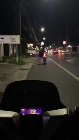This is a first-person perspective video taken from a motorcycle at night. The rider is following another motorcycle with a person on the back. The road appears to be a city street with buildings, streetlights, and other vehicles. A sign in Thai and English reads "HOMA". The dashboard of the motorcycle shows a speed of 14 km/h. The scene is dark, illuminated by streetlights and vehicle lights.