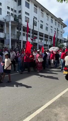 The video captures a daytime scene on a street in Delkanda, Sri Lanka, showing a large group of people marching. Many individuals are dressed in red clothing and are carrying numerous red flags on poles, suggesting an organized rally, protest, or procession. They are walking along a paved road from left to right across the frame.

The weather appears sunny and clear, with strong light casting distinct shadows on the ground. A silver Suzuki Alto hatchback, bearing the Sri Lankan license plate "SP CAN 1946," is visible in the foreground, either parked or slowly navigating alongside the marchers.

A large, multi-story white building with numerous windows and balconies lines the street, forming the primary backdrop for the procession. Utility poles and overhead wires are also present. Some small signs are affixed to the white building, with one partially visible as "HIGHLAND." Green foliage from trees is visible in the upper right corner of the frame, adding a touch of nature to the urban setting. The event appears to be a public gathering in an urban area.