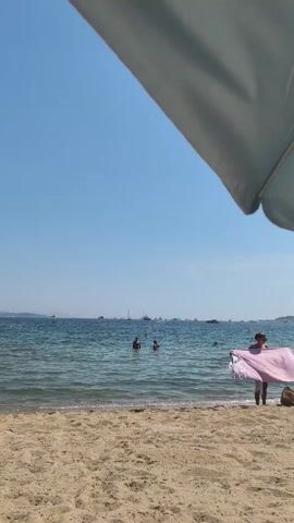 The video shows a beach scene with a sandy shore in the foreground. The ocean is calm with gentle waves. Several people are in the water, some swimming and others standing. In the right foreground, a person is standing near the water's edge holding a pink towel. In the distance, there are several boats on the horizon under a clear blue sky. A large umbrella shades part of the foreground.
