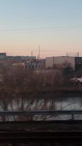 The video shows a landscape with industrial buildings and a river, seen from a moving vehicle. The buildings are primarily white and gray, varying in size and shape. A few bare trees stand in front of the buildings, and power lines stretch across the sky. The river reflects the buildings and the sky, which has a gradient of light blue and peach hues, suggesting either sunrise or sunset. A metal guardrail is visible in the foreground, indicating the perspective is from a bridge or road. The scene appears to be taken during the late afternoon or early morning when the light is soft and diffused. There are no people visible.