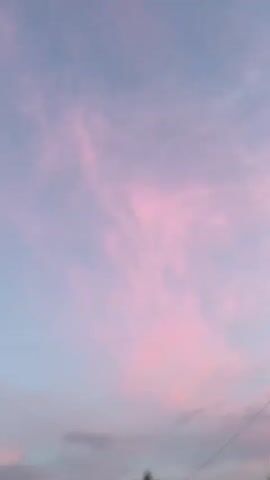 A video shows a time-lapse view of the sky at sunset. The sky transitions from light blue with pink clouds to a vibrant orange and red as the sun sets. Silhouetted trees and buildings are visible in the foreground. Power lines are also visible across the sky.