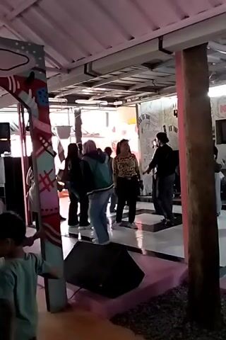 A group of people are dancing in a covered outdoor area. There is a stage with speakers, and a microphone stand. The walls are decorated with colorful artwork and some text. The floor is paved. There are pillars supporting the roof, one of which is wrapped in a colorful fabric. Some of the people are wearing casual clothing, and some are wearing headscarves. The atmosphere appears festive.