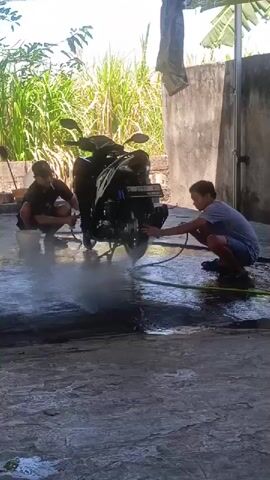 TWO YOUNG BOYS ARE WASHING A BLACK MOTORCYCLE OUTSIDE. THE BOY ON THE RIGHT IS HOLDING A GREEN HOSE AND SPRAYING WATER ON THE REAR TIRE OF THE MOTORCYCLE. THE BOY ON THE LEFT IS HOLDING A WHITE BUCKET AND APPEARS TO BE HOLDING IT UNDER THE REAR WHEEL OF THE MOTORCYCLE. WATER IS SPLASHING ON THE CONCRETE GROUND. THE BACKGROUND CONSISTS OF TALL GREEN GRASS AND A CONCRETE WALL. THE SCENE IS LIT BY BRIGHT SUNLIGHT.