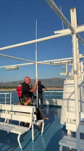 The video shows a group of people on the deck of a white ferry with blue flooring. The sky is clear and blue, and the sea is visible in the background, with a distant coastline and mountains. Several people are standing near the railing, looking out at the sea. One man in a black shirt is gesturing towards the sea, possibly pointing something out to others. A woman with a black top and skirt is seated on a white bench. Other individuals are visible in the background, also observing the scenery. The ferry appears to be in motion, as indicated by the slight blur of the water and distant objects.