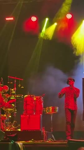 A man in a patterned shirt, black pants, and a hat is singing into a microphone on a stage. He is standing to the right of a drum set. The stage is lit with red and yellow spotlights, and there is smoke in the air.