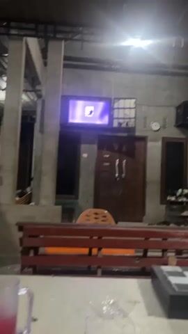 The video shows a television screen displaying a purple background with a silhouette of a woman. The television is mounted on a grey concrete wall above a wooden door. To the left of the door is a window, and to the right is another window. In front of the television and door is a reddish-brown wooden bench. There is also an orange chair behind the bench. The scene appears to be outdoors at night, with artificial lighting illuminating the area. The overall environment seems to be a residential setting.