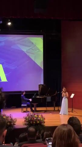 A full shot captures a singer and pianist on a stage performing for an audience. The singer, a woman with blonde hair, stands to the right of the stage, wearing a long, light blue gown. She holds a microphone to her mouth, actively singing. To her left, a man in a dark suit sits at a black grand piano, playing. Behind them, a large screen displays a purple and green abstract design with a large yellow "A" visible on the left side. The stage is adorned with floral arrangements in front of the performers. The audience is seated in the foreground, mostly out of focus. The lighting suggests a formal performance setting.