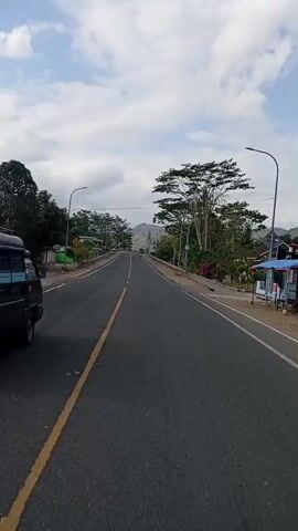 The video shows a view from a vehicle driving on a road. The road is paved and has a yellow line dividing the lanes. There are trees and buildings on the sides of the road. The sky is cloudy. A black pickup truck is visible in the distance, and a motorcycle is seen passing by. The camera appears to be mounted on the front of the vehicle.