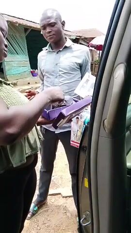 A woman standing beside an open car door examines the contents of a purple box handed to her by a man standing in front of her. The man is holding a box of two white bottles. The background shows street-side businesses. The woman is wearing a light green top and black pants. The man is wearing a light grey button-up shirt and dark pants.  The scene appears to be outdoors in daylight.