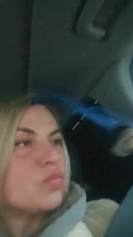 A woman with blonde hair sits in a car and holds up a pair of glasses with a black frame and blue temples. She makes a kissing face at the camera. The interior of the car is visible, including the ceiling, a headrest, and a window covered by a dark shade.
