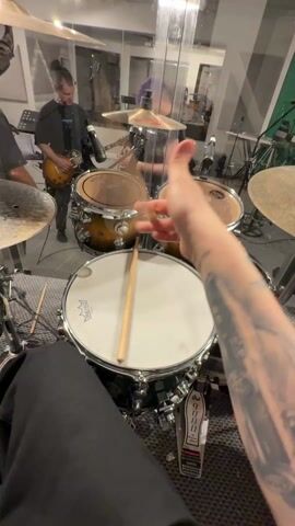 The video shows a first-person view of someone playing a drum set. The person is holding drumsticks and preparing to play. The drum set includes a snare drum, two tom-toms, a cymbal, and a bass drum pedal. The person is wearing black pants and has a tattoo on their left arm. In the background, a man plays an electric guitar, and another person stands near a microphone. The scene appears to be a music studio or practice room. The background includes a glass barrier, other instruments, music stands, and studio equipment.