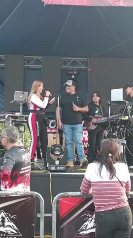 A group of musicians are performing on an outdoor stage. A woman in a white, black, and red jumpsuit speaks into a microphone to the left. A man in a black t-shirt, jeans, and a baseball cap stands next to her, holding a microphone. Behind them, there is a young man playing a guitar, a man working on a laptop, and another man to the right. There is a backdrop of black LED screens and a metal framework. There are a few people in the audience watching the performance, with the back of a woman in a striped shirt and black jeans visible prominently in the foreground. There are red, white, and black banners at the front of the stage. The setting appears to be a public outdoor event, possibly a festival or fair.