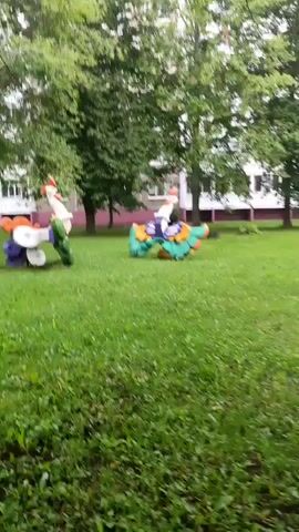 Two colorful, abstract sculptures resembling birds or mythical creatures are positioned on a grassy lawn. The sculpture on the left is primarily white, purple, and orange, with green accents. The sculpture on the right is predominantly green, yellow, blue, and purple. Both sculptures appear to be made of recycled materials, possibly plastic. They are situated in an outdoor setting with trees and a building in the background. The grass is green and appears well-maintained. The lighting suggests an overcast day.