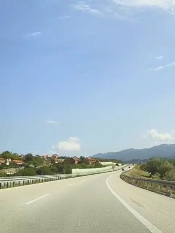 The video captures a view from a moving vehicle on a highway. The road ahead curves to the right, with a guardrail separating the lanes. To the left of the highway, there are houses with red roofs nestled amongst green trees and vegetation. In the distance, mountains are visible under a clear blue sky with scattered white clouds. The perspective suggests the vehicle is traveling on the right side of the road. There are no people or other vehicles immediately visible in the frame. The environment appears to be a rural or semi-rural setting.