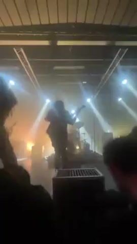 A band performs on a lit stage in front of a crowd, captured from the crowd's perspective. The stage lighting is bright, with beams illuminating the performers. The lead singer, silhouetted against the lights, engages with the audience. Other band members are visible behind him. The crowd is visible in the foreground, with hands raised. The venue appears to be an indoor space with a high ceiling and exposed structural elements. The video has a slightly shaky quality, likely due to the movement of the person filming.