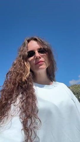 A medium shot video shows a woman with long, curly brown hair and sunglasses looking at the camera. She is wearing a light blue sweatshirt. The background shows a bright blue sky and a rocky, uneven terrain with sparse green vegetation. The video is shot outdoors on a sunny day.