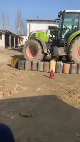 The video shows a green Claas tractor parked in a dirt yard in front of a white building. A row of various colored jerry cans is positioned in front of the tractor’s front left tire. A man wearing a black turtleneck and brown pants walks into the frame and stands near the rear tire. He looks at the camera and adjusts his clothing. The background includes a building, trees without leaves, and various metal objects on the ground.