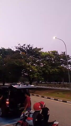 A group of people are gathered near a black SUV with its trunk open. Two individuals in red helmets are on a motorcycle to the right of the SUV. The sky is a light purple and there are trees in the background. Some birds are flying in the distance.