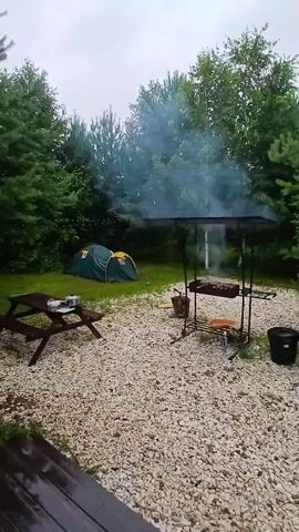 The image captures a campsite scene with a green tent pitched on a grassy area, surrounded by dense trees. A picnic table with plates sits to the left, while a barbecue grill with food cooking on it is centrally located. Smoke billows from the grill, suggesting an outdoor cooking activity. The ground is covered in light-colored gravel. The weather appears overcast, with a muted, diffused light typical of a cloudy day. The overall atmosphere is one of a relaxed outdoor gathering, possibly a camping trip or a barbecue. There are no visible people in the image, but their presence is implied by the setup. The time of day is not clearly indicated, but the lighting suggests it could be morning or afternoon. The location cues of trees and a natural outdoor setting are consistent with a campsite in a wooded area.