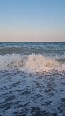 The video shows a close-up of ocean waves crashing on a dark-colored beach under a clear blue sky. The waves are foamy and white as they break and recede. The sun appears to be low in the sky, casting a warm glow on the water. There are no people or objects visible in the frame.