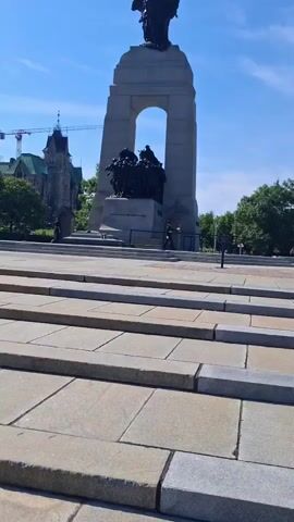 The video shows a monument in front of a building with a crane in the background. The monument is a large stone structure with sculptures. There are steps leading up to the monument. There are two people in uniform standing at the base of the monument on either side. The sky is blue with a few clouds. The day is sunny.